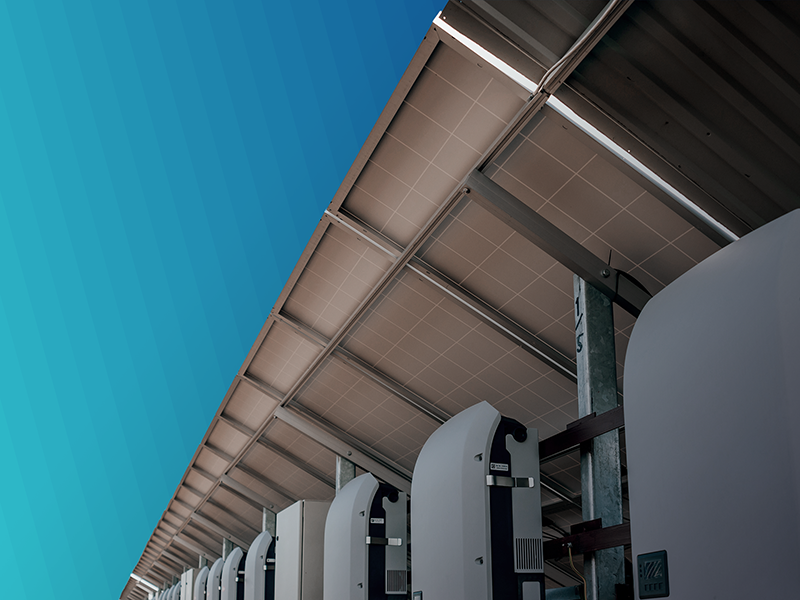Row of solar inverters mounted beneath a large solar panel array against a turquoise gradient background.