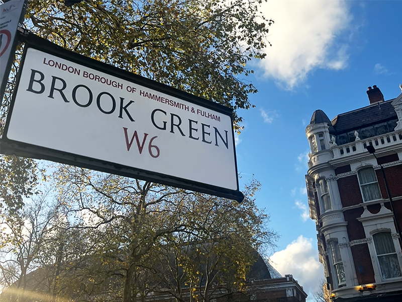 A close up of a sign saying Brook Green W6, backed by trees, a grand Victorian building a blue sky littered with white clouds.