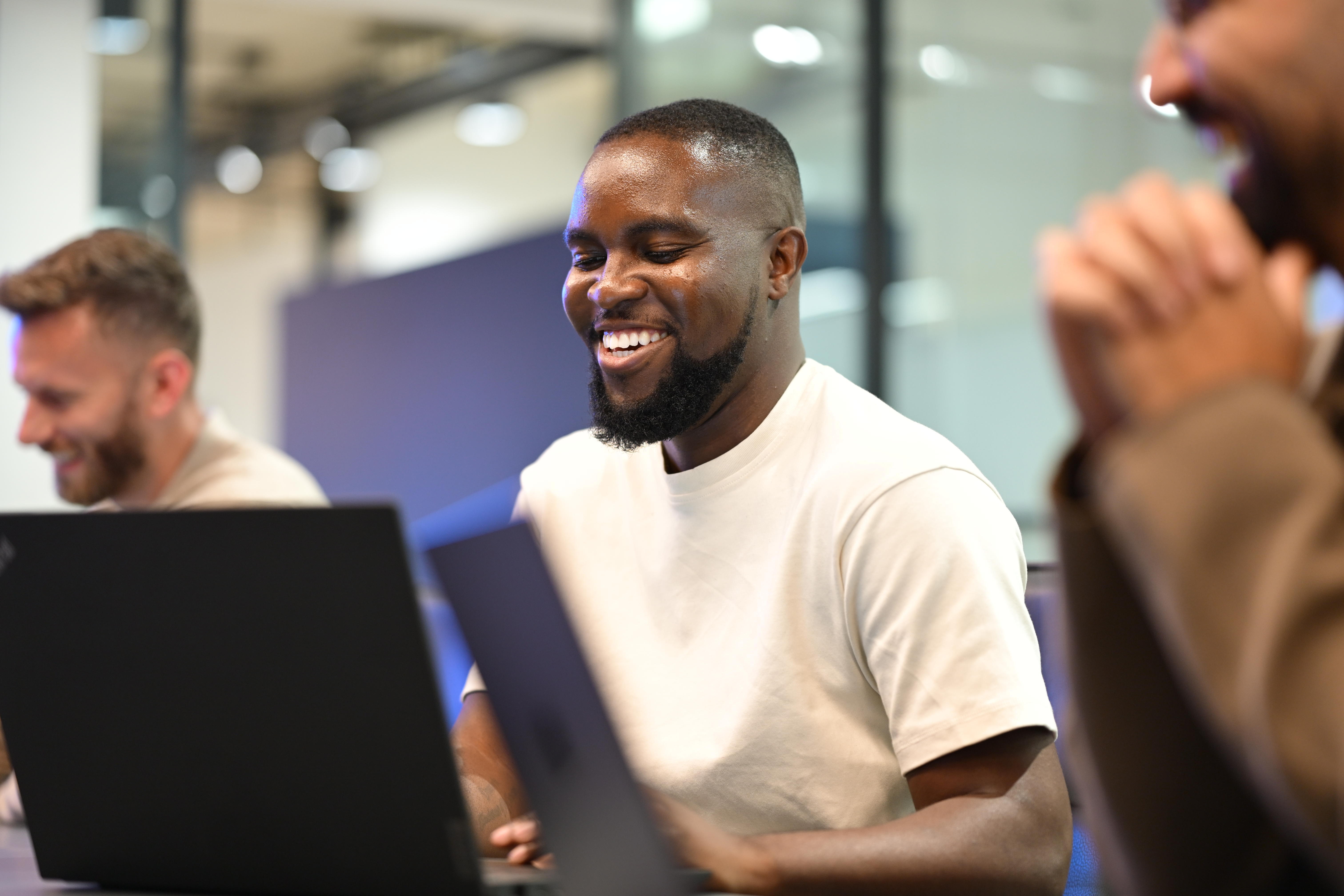 Three people sat at a table with laptops smile and laugh.