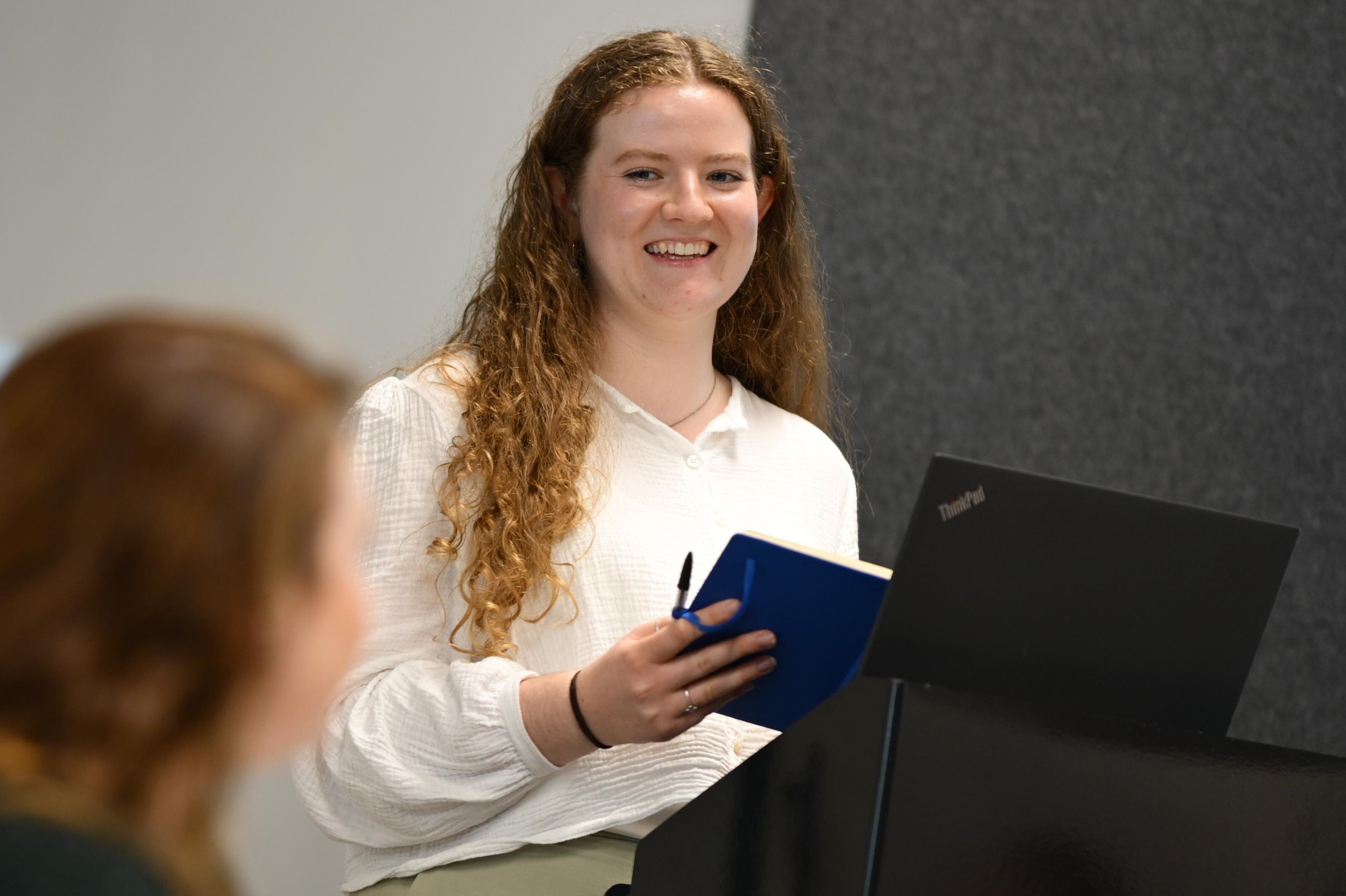 A person stood at a podium smiles as they look at a person sat down to their side.
