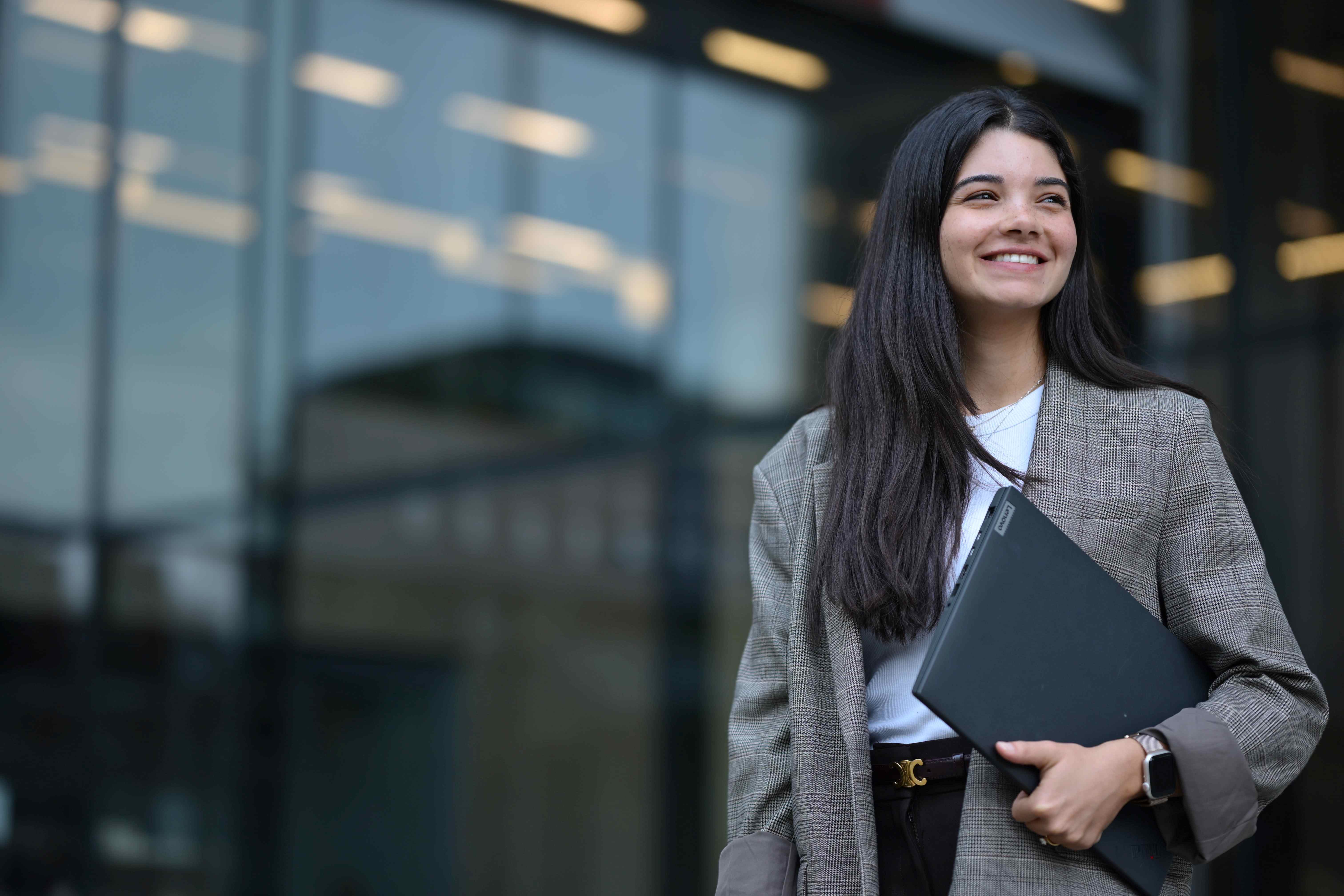 A person standing outside an office building with a laptop smiles as they look off to the side.