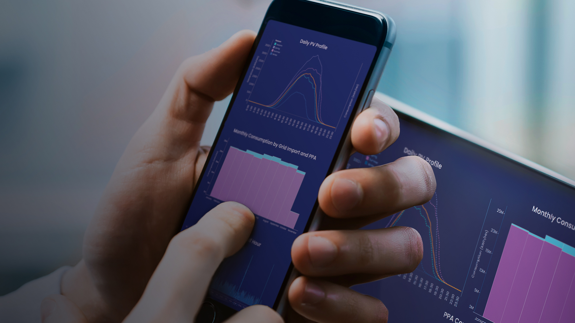 Hand holding a smartphone displaying energy-usage data, with a laptop in the background showing similar analytics.