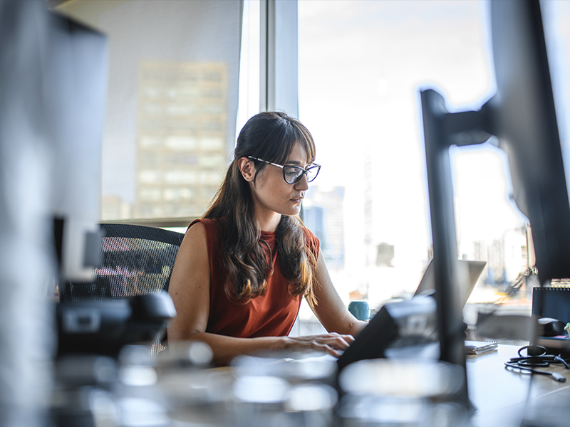 A side view of a person on a laptop in an office environment surrounded by computer equipment.
