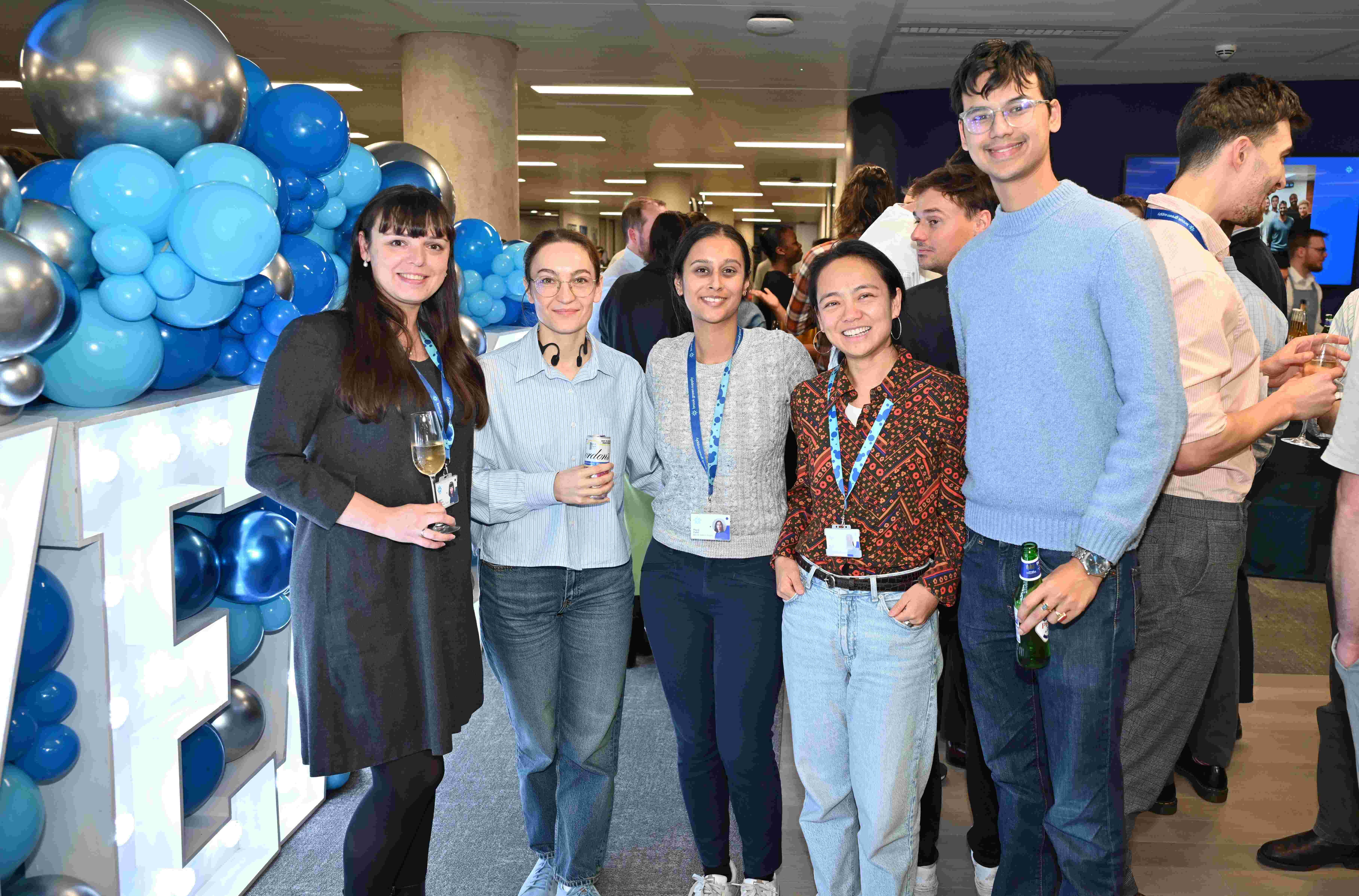 A group of people pose for the camera with drinks, with balloons and light up letters in the background