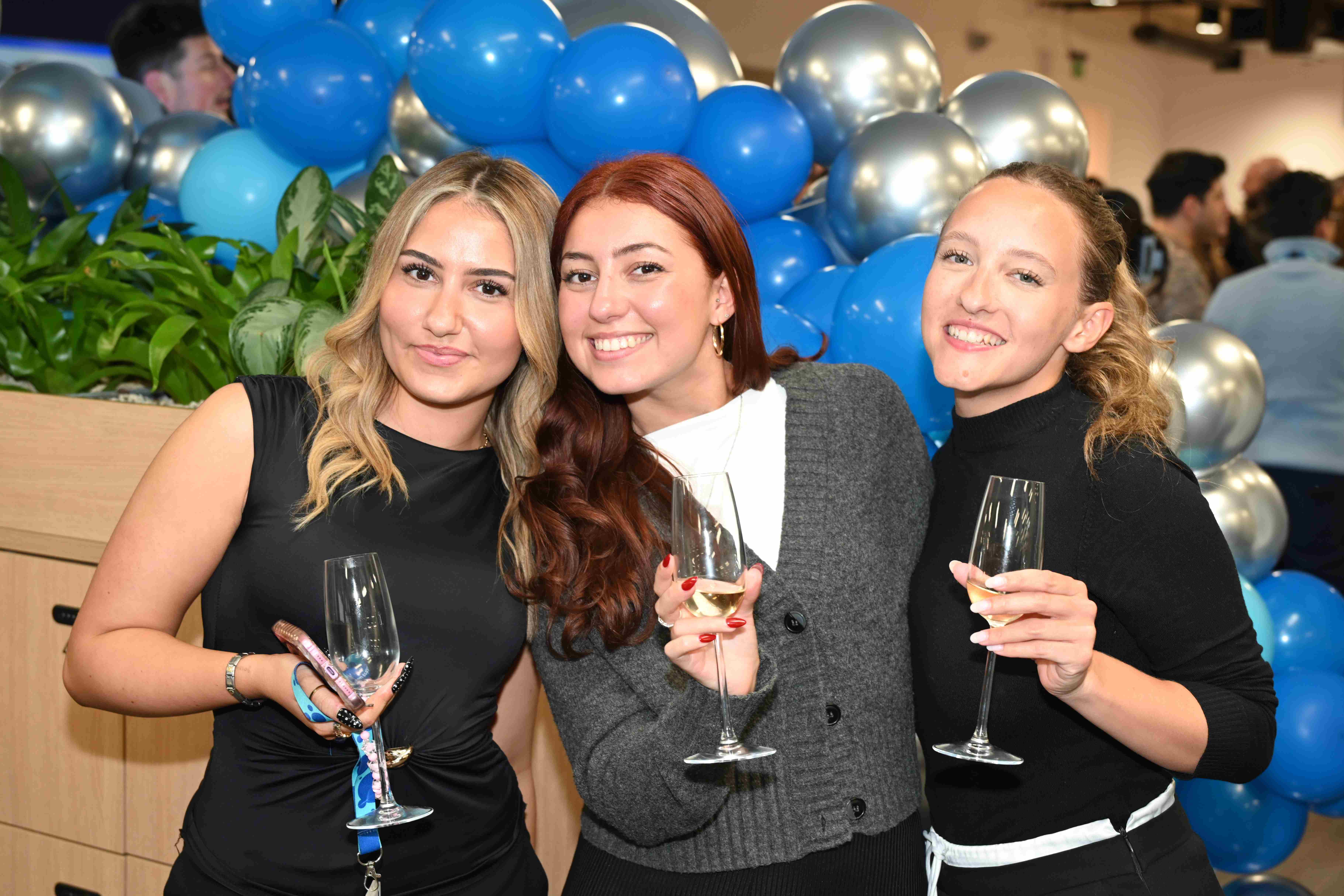Three people smile and pose with drinks, with blue and silver balloons in the background