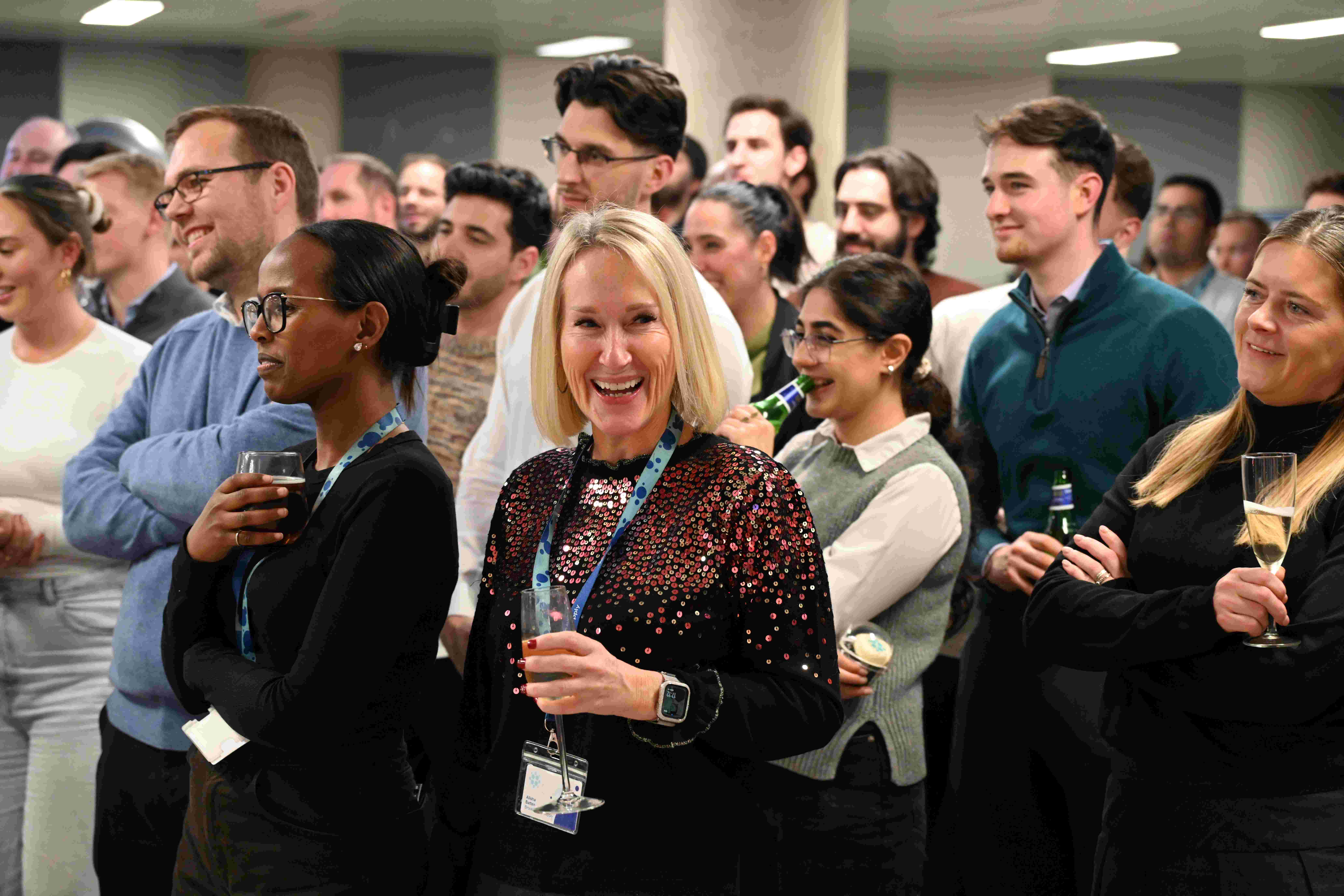 A crowd of people watch a video and smile, with drinks in their hands
