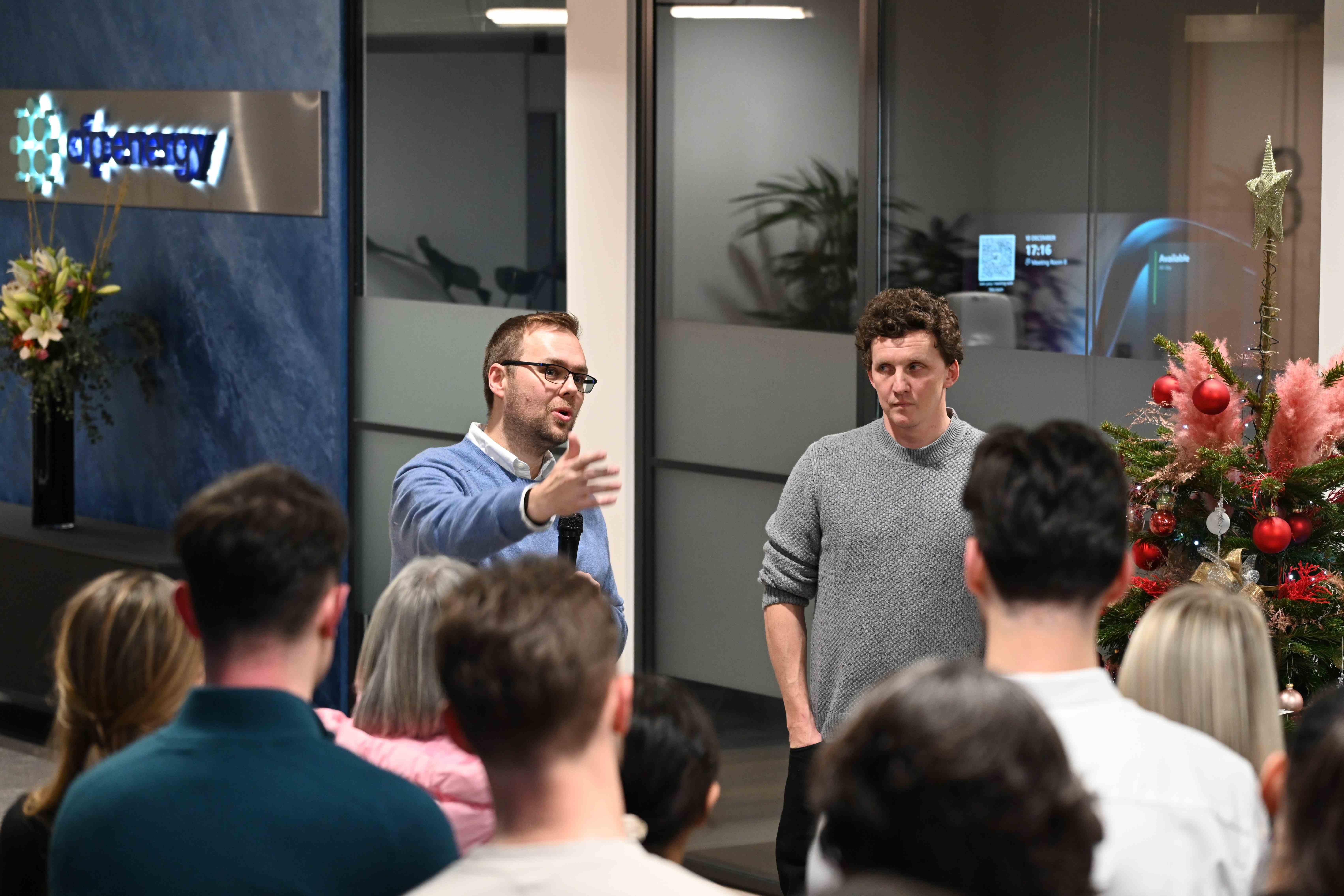 Two people stand in front of a crowd. One person speaks into a microphone