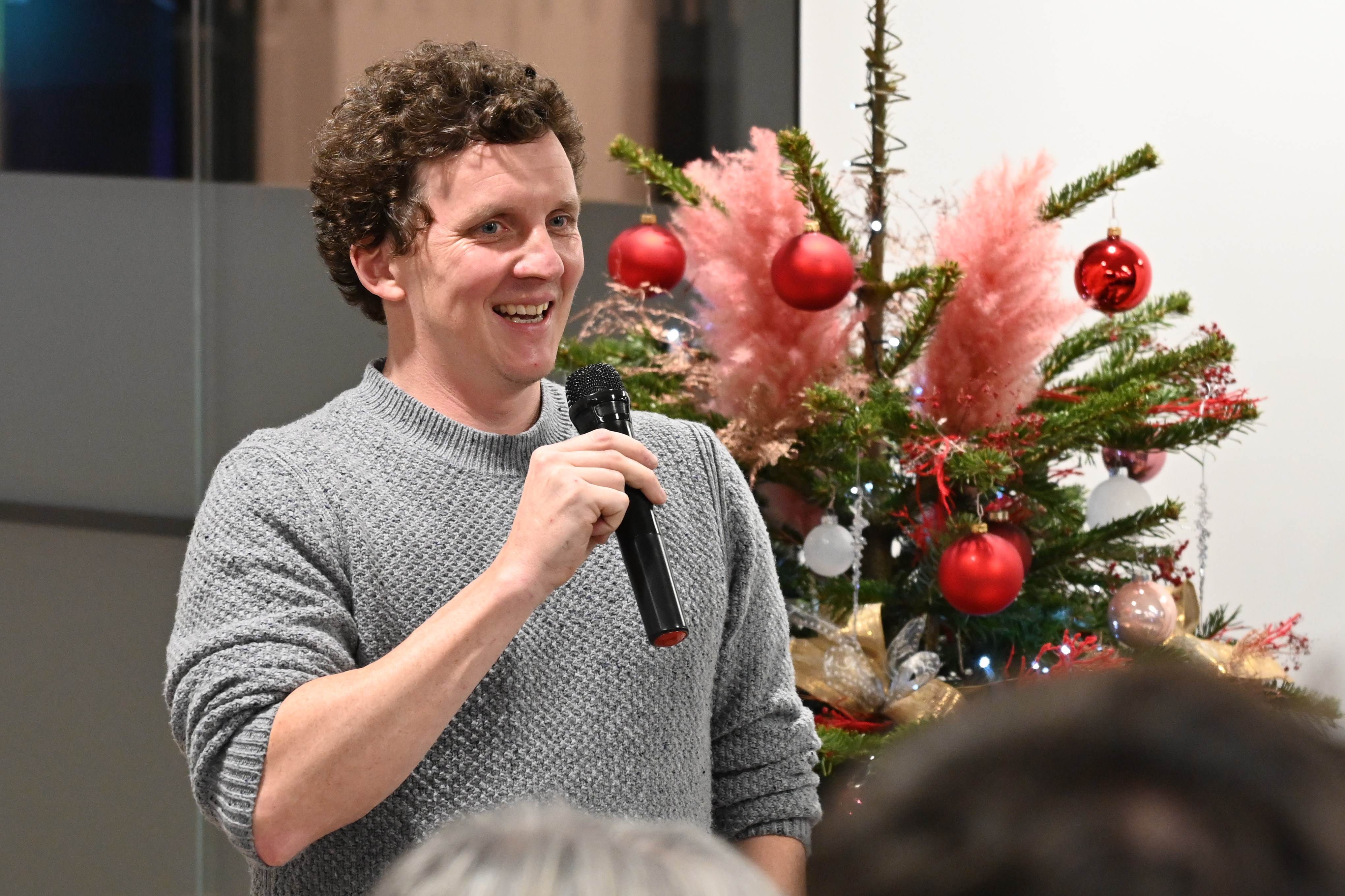 A person talks into a microphone in front of a Christmas tree