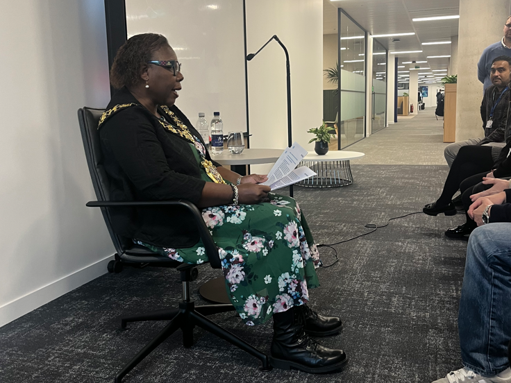 A side view of a person reading from some notes in front of a crowd in an office.