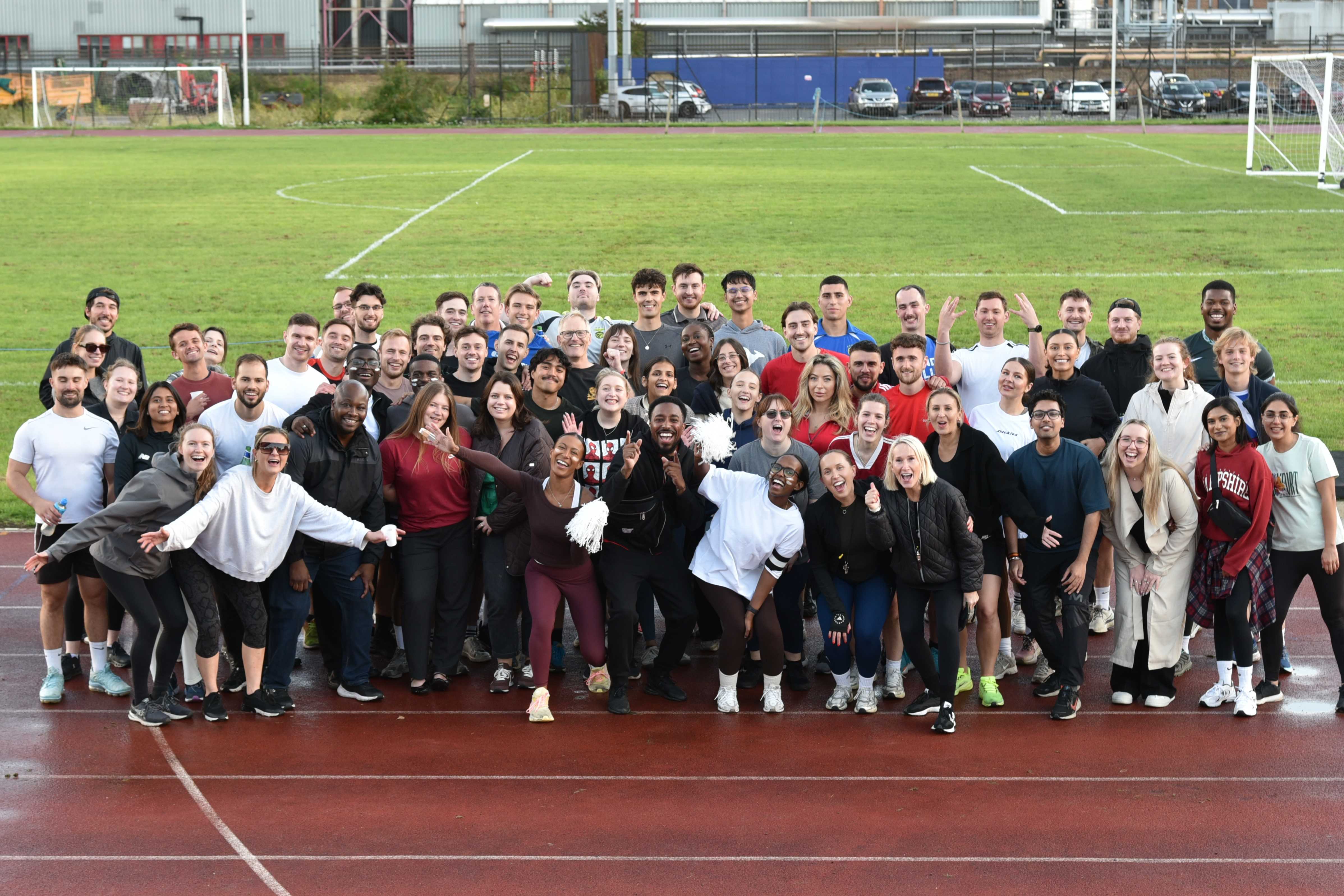A large group of people dressed in sports clothes smile while stood on a sports track.