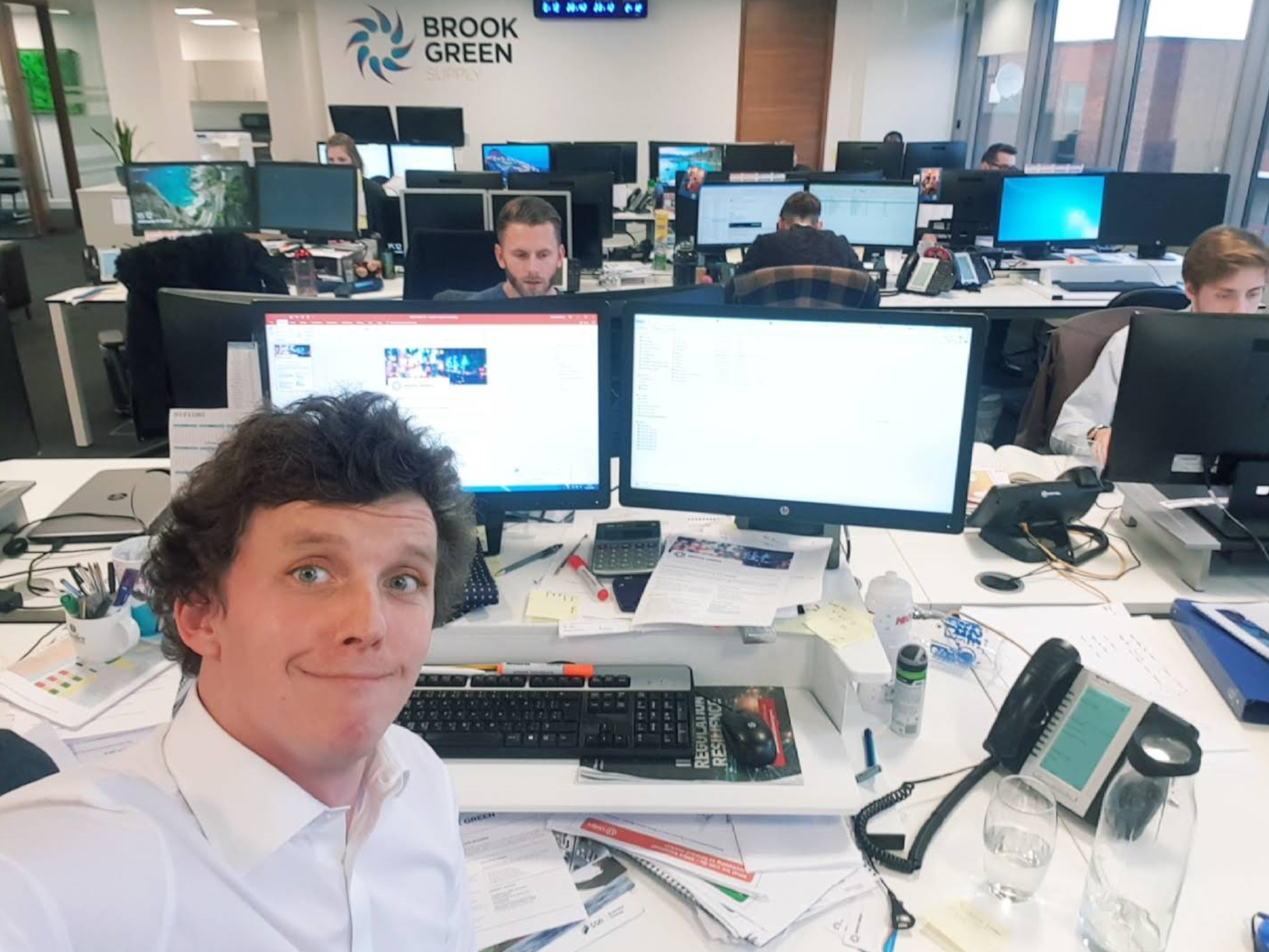 A person at the front of a busy open-plan office takes a selfie while seated at a cluttered desk with multiple monitors, papers, and office supplies. Several colleagues work at their computers in the background, and a wall sign reads ‘Brook Green Supply'.