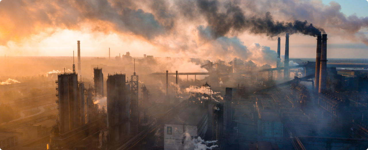 A wide landscape of a large factory that's emitting large amounts of smoke, set against a setting sun.