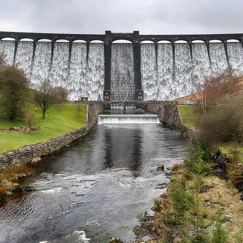 A wide view of a hydro dam with water cascading down its face and flowing into a tree-lined river.
