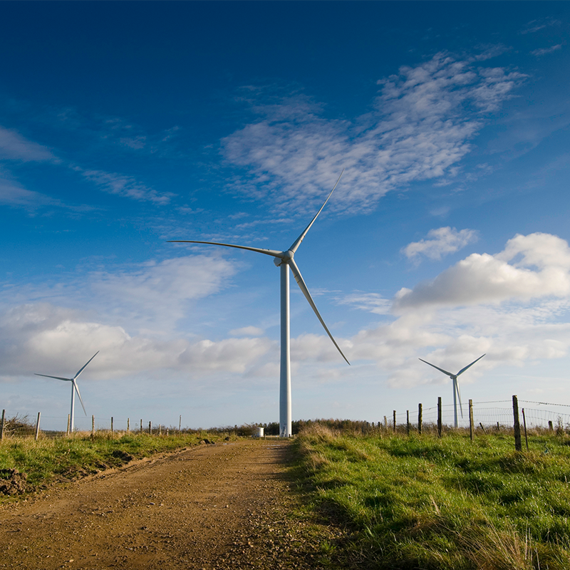 Three wind turbines in a row with one in the foreground, set against a blue sky with light white cloud.