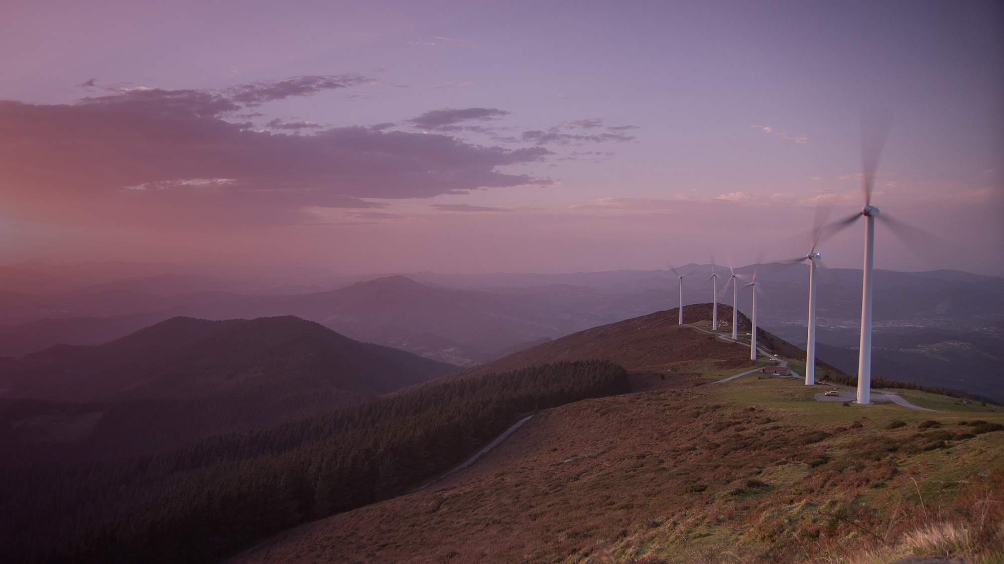 A row of wind turbines atop a hill set against a glowing pink sky.
