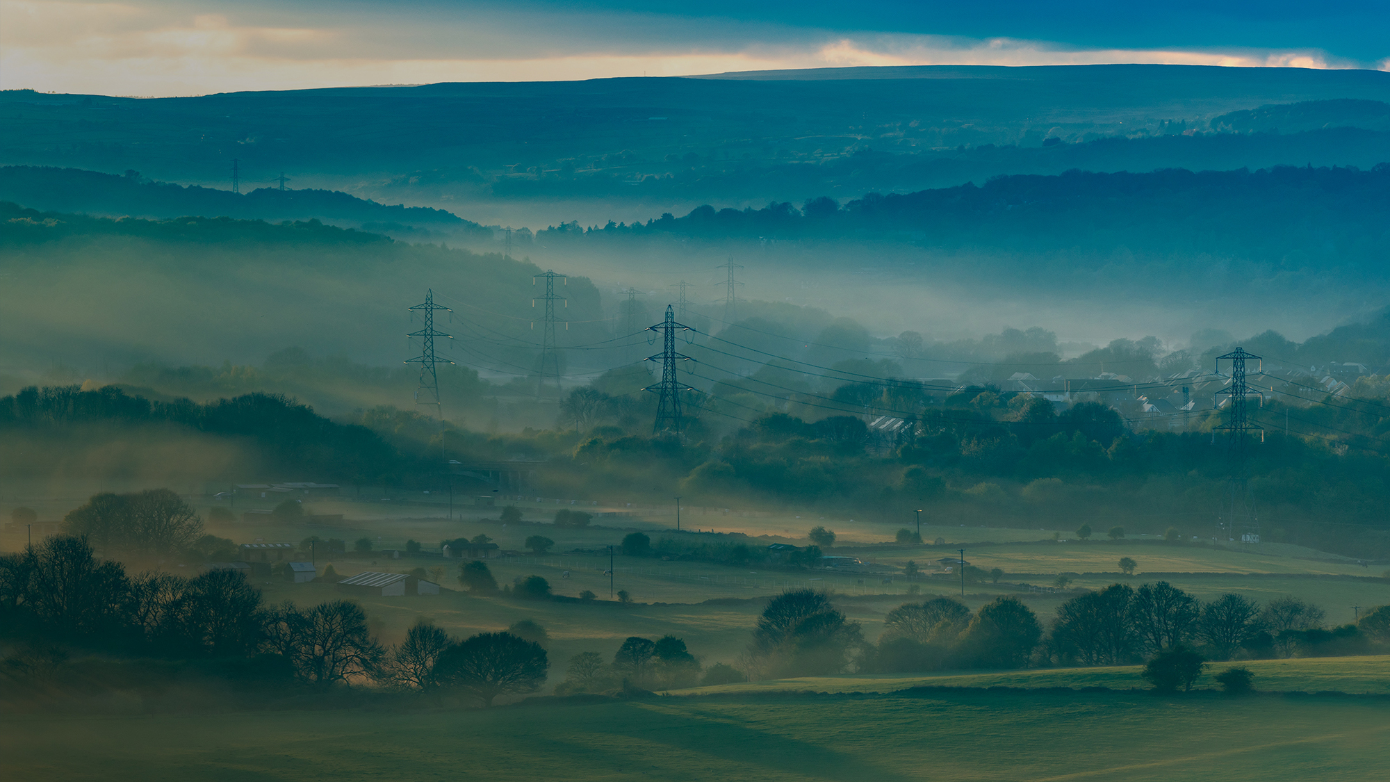 A wide view of a sequence of electricity pylons set amidst a misty landscape of green fields and trees.