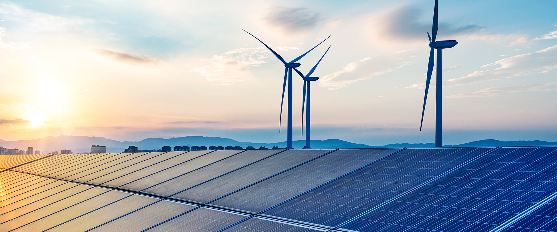 A row of solar panels with wind turbines in the background, set against some low hills with a hazy sky.