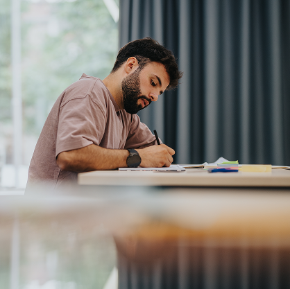 A side view of a person sat at a table scribbling on a notepad.