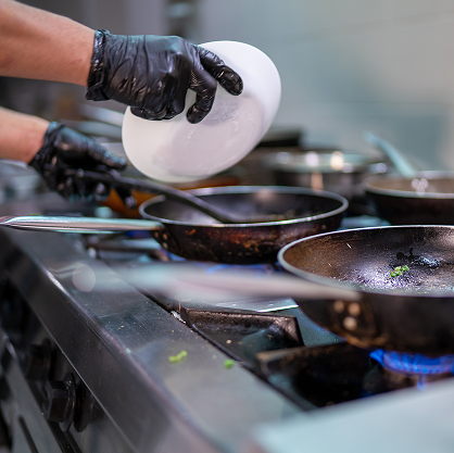 Close up of two black gloved hands spooning something from a sizzling frying pan into a bowl.