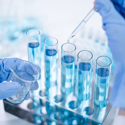 Blue gloved hands drop something from a pipette into a row of test tubes full of blue liquid.