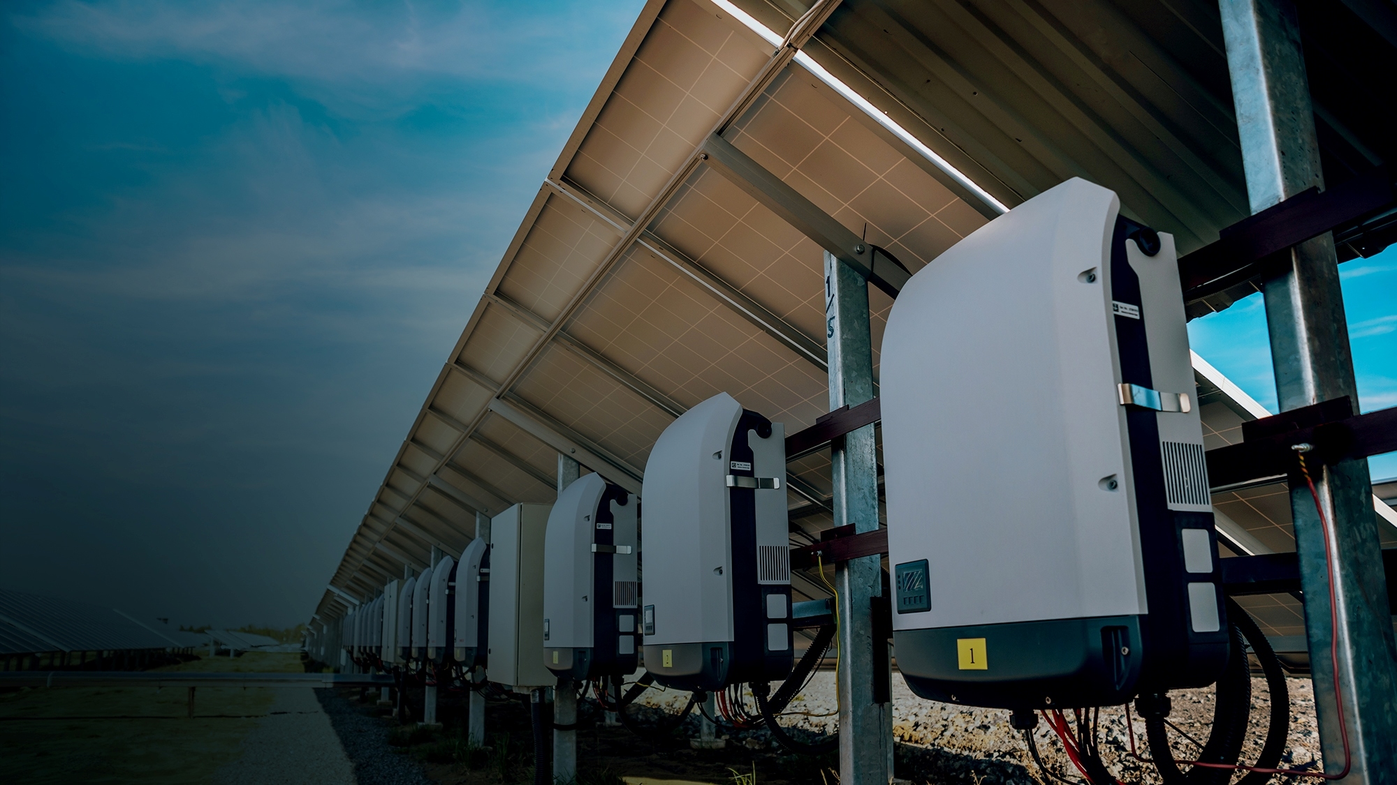 Row of solar inverters mounted beneath a large solar panel array outdoors, with blue sky in the background.