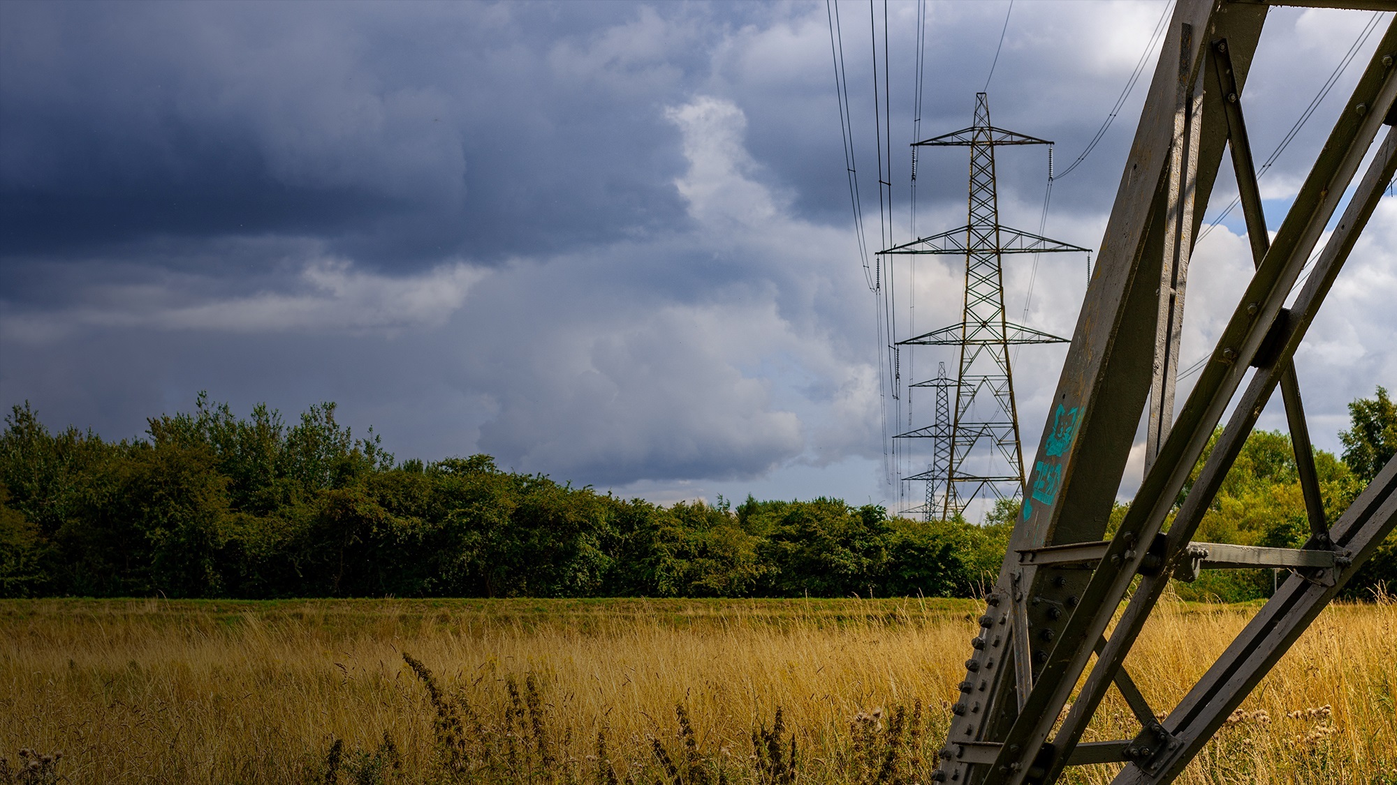 A close up of the foot of an energy pylon in a field, backed by further energy pylons that recede into the background against a cloudy sky.