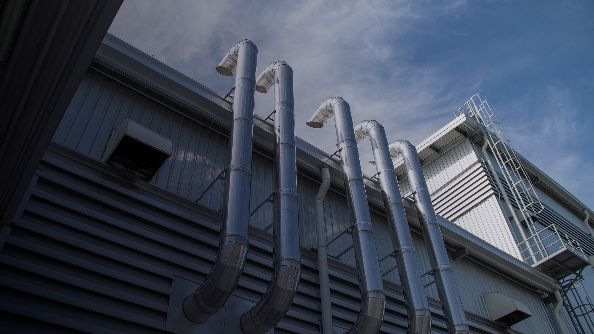 Exterior view of an industrial building with multiple large, shiny metal ventilation pipes running vertically along the wall against a partly cloudy sky.